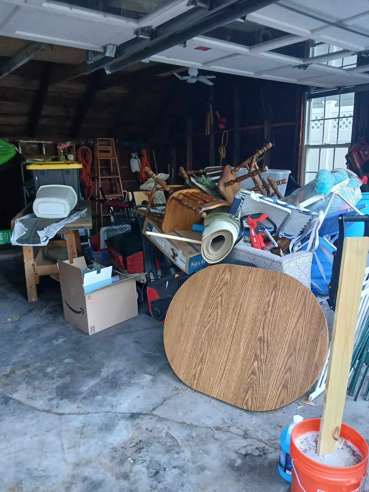 A garage piled along one side with a table top, stacked chairs, a rolled carpet, boxes and lawn chairs